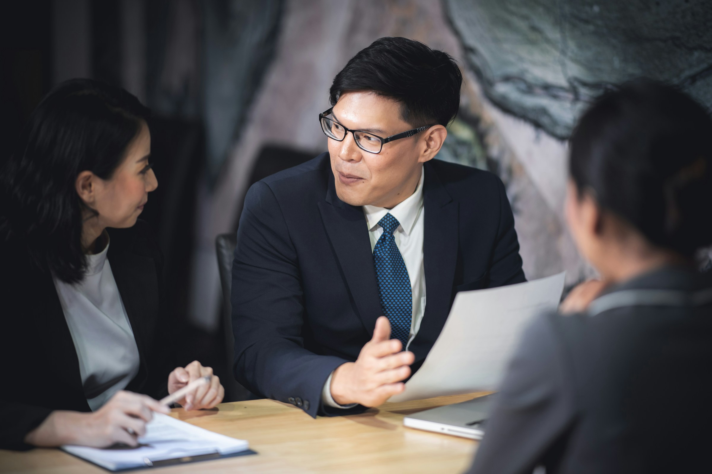 Three business professionals in a discussion, with one man in glasses holding a paper and gesturing at a wooden table.
